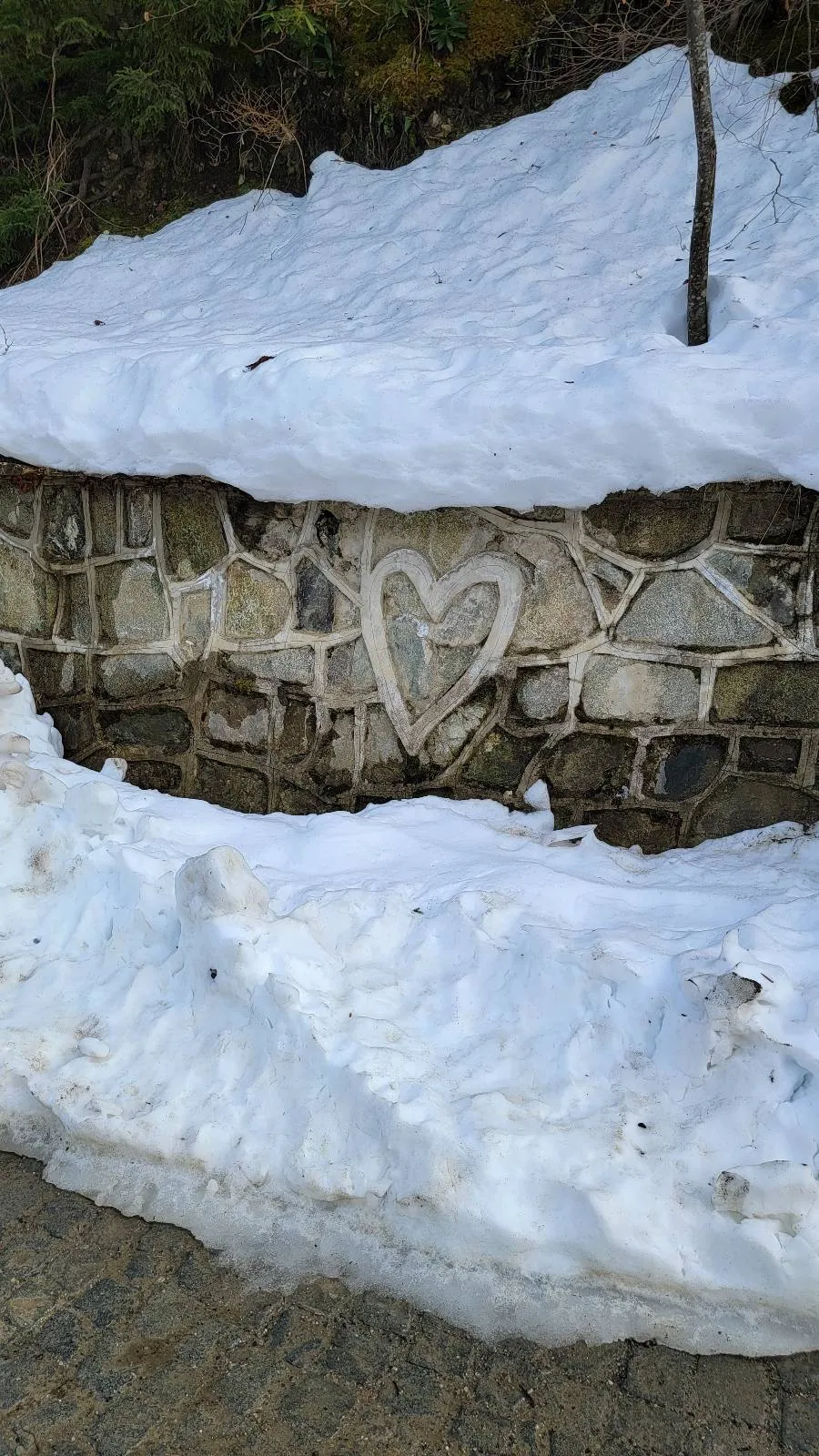 A stone wall with a heart carved into it is partially covered by snow.