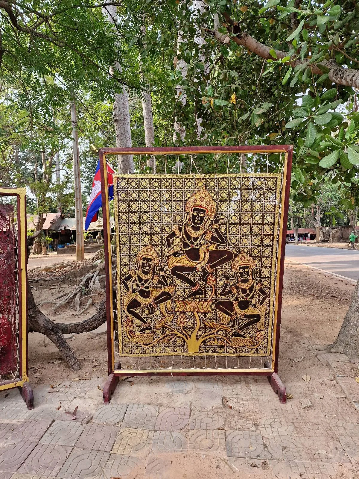 A decorative metal panel with intricate golden designs, featuring three figures in traditional attire. It's displayed outdoors on a stand, with lush green trees and a flag in the background.