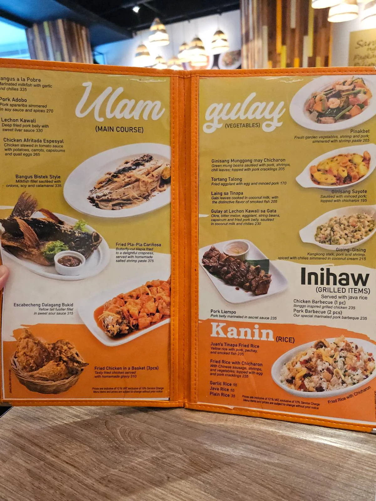 A photo of a restaurant menu featuring various dishes. The left page lists "Ulam" with images of meat and seafood dishes. The right page includes "Gulay," "Inihaw," and "Kanin," showing vegetables, grilled foods, and rice dishes.
