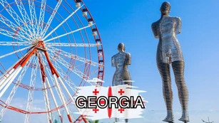 A large Ferris wheel stands beside two tall, segmented metal statues of human figures under a clear blue sky, with the word "GEORGIA" and the Georgian flag in the foreground.
