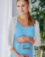 A pregnant woman stands indoors, gently holding her belly while wearing a blue shirt with the words "baby on board."