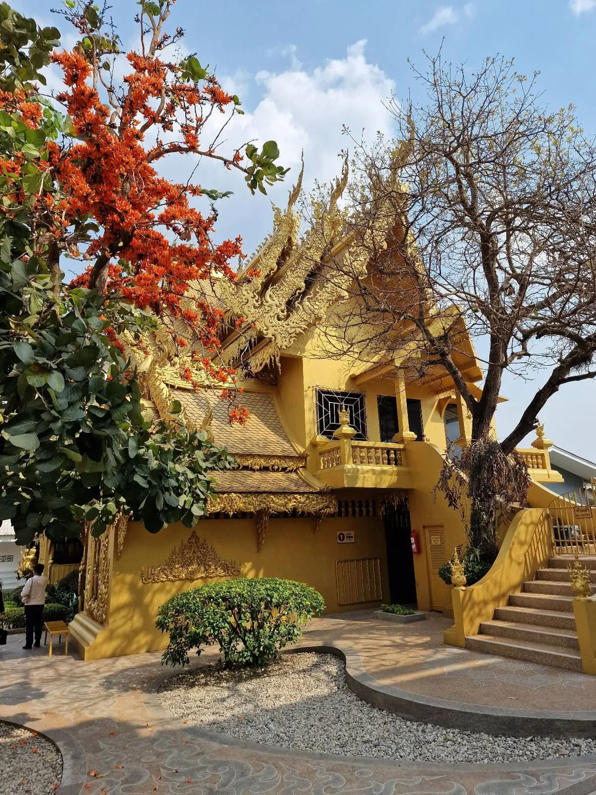 A golden temple with intricate details under a blue sky, surrounded by trees. A prominent tree with orange flowers stands in the foreground, and a staircase leads up to the entrance.