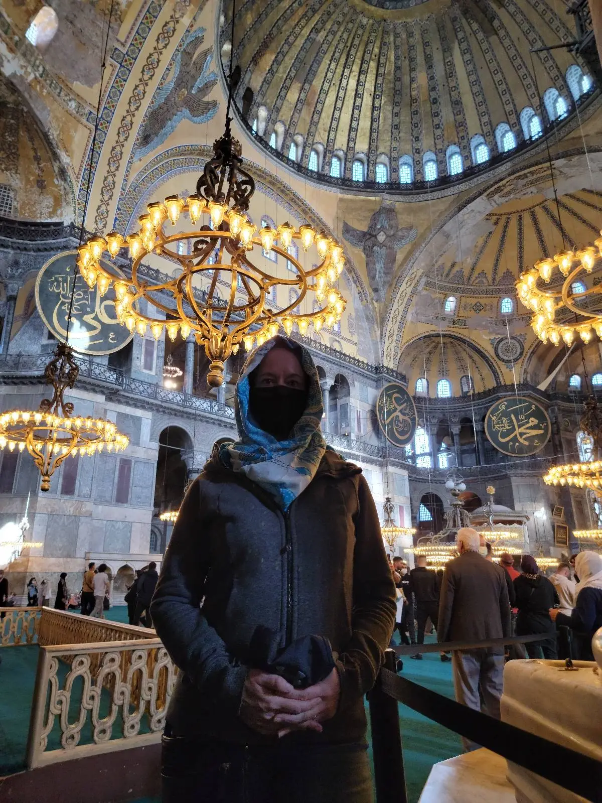 A person wearing a headscarf stands inside a grand, ornate building with large chandeliers and decorative ceilings.