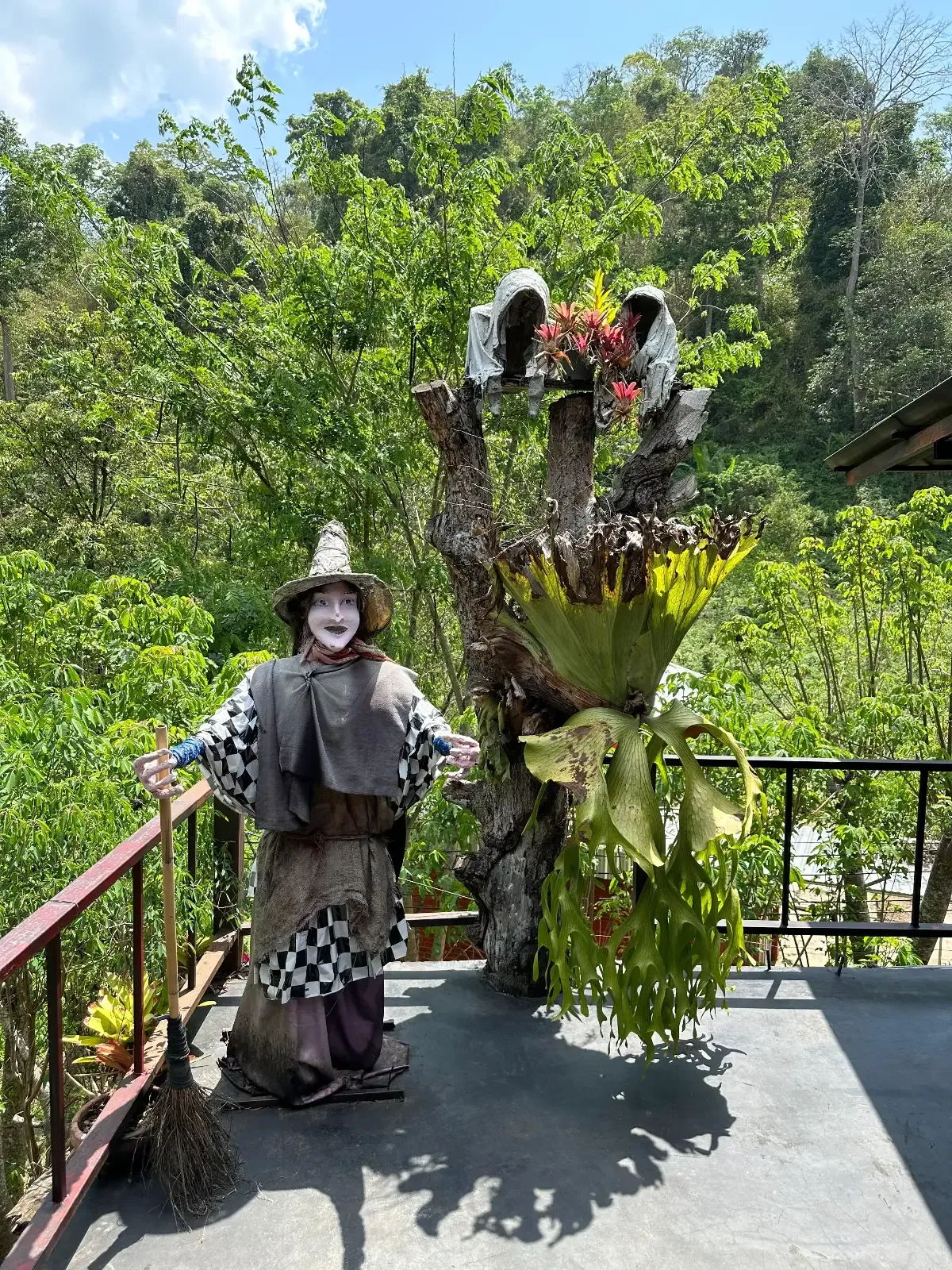 A person in a black-and-white clown costume and makeup stands on a balcony next to a large, tree-like costume with greenery, against a backdrop of lush green forest and clear sky.