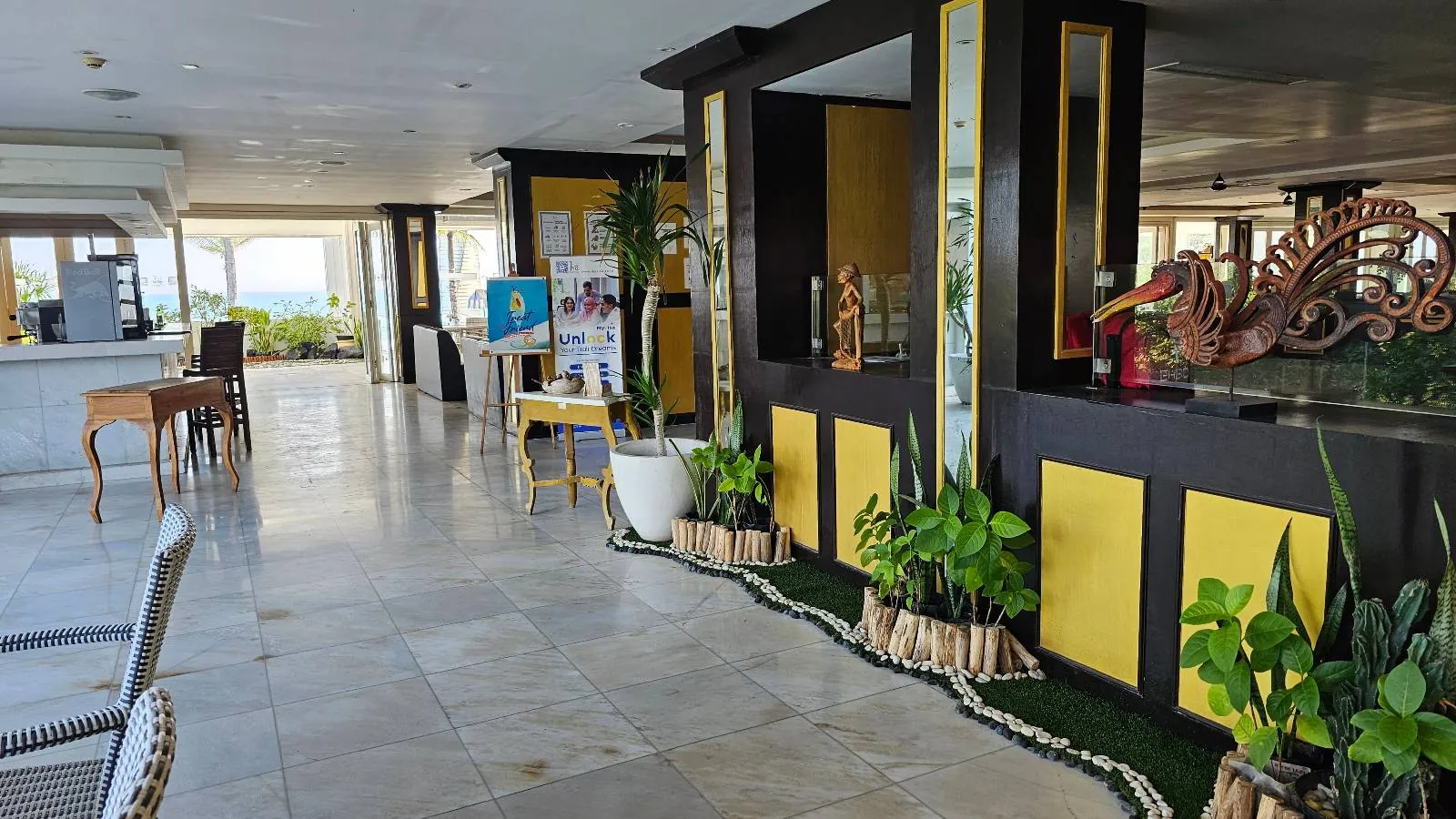 Lobby area with marble flooring and yellow and black decor. Green plants line a dark partition, and there's a wooden desk nearby. Art pieces and tropical scenery through windows create a welcoming, bright atmosphere.