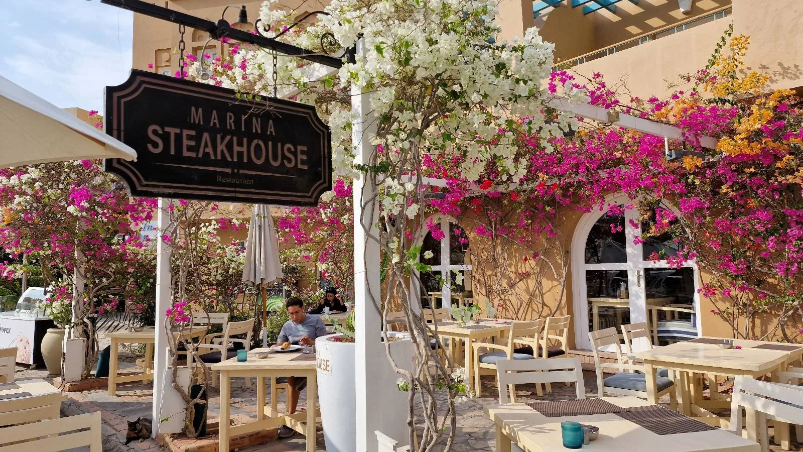 Outdoor seating area of Maizon Steakhouse, adorned with blooming pink and white flowers, wooden tables, and chairs beside a light-colored building with arched windows.