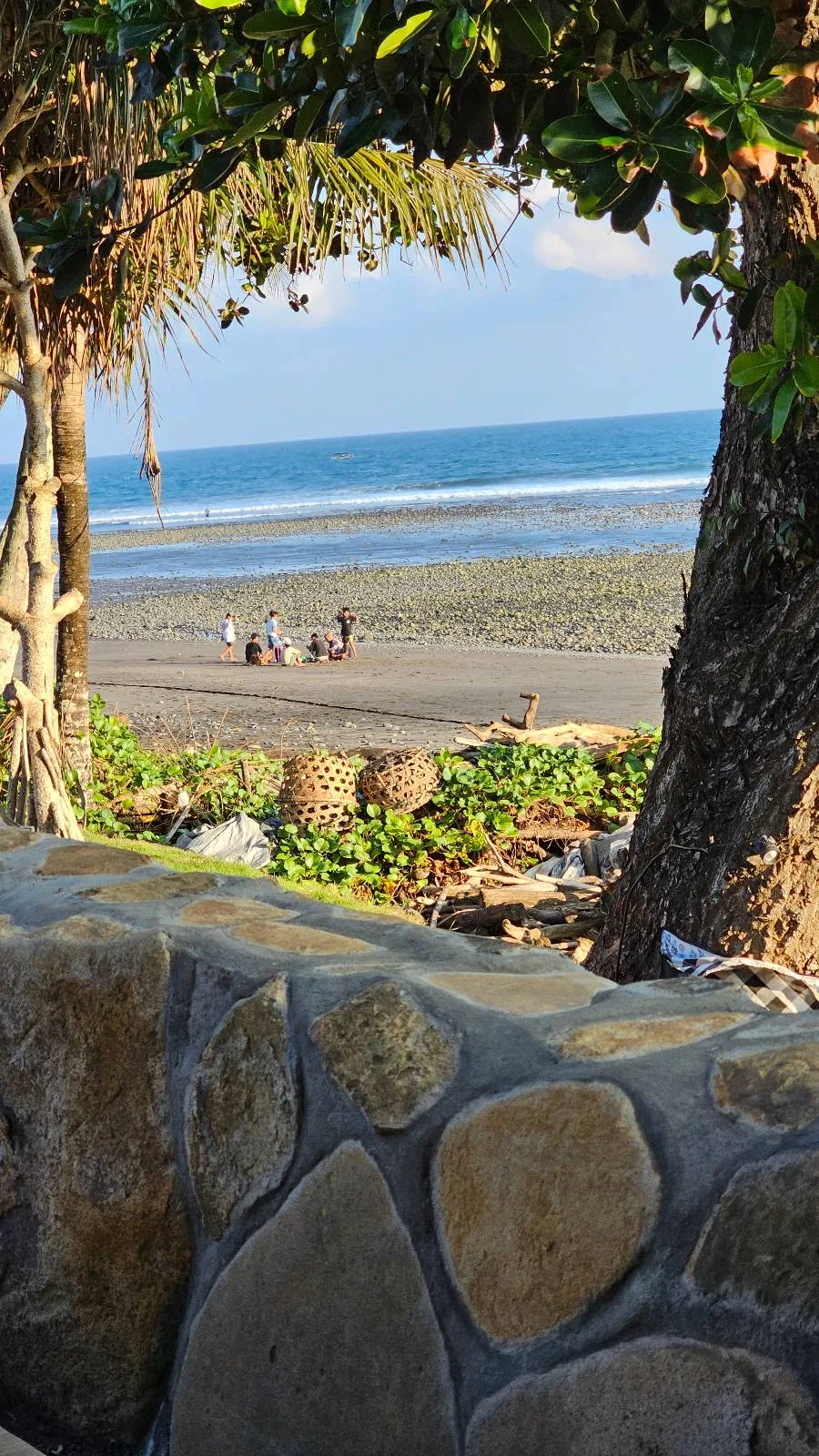 View of a beach through trees, featuring a stone wall in the foreground. People are sitting on the sand near the shoreline. The ocean is calm with gentle waves under a clear blue sky. Greenery surrounds the area, adding a tropical feel.