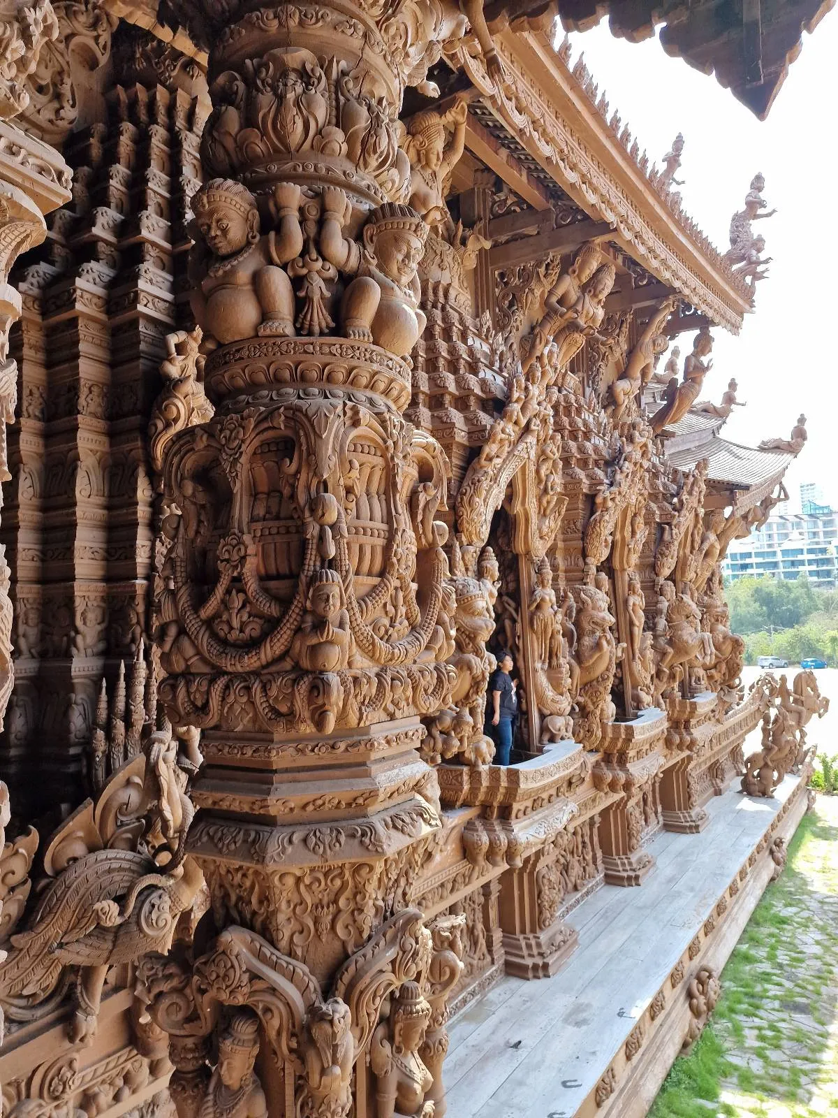 Detailed carvings on ornate wooden pillars and walls of a temple, featuring intricate patterns and figures. Sunlight highlights the craftsmanship against a backdrop of greenery.