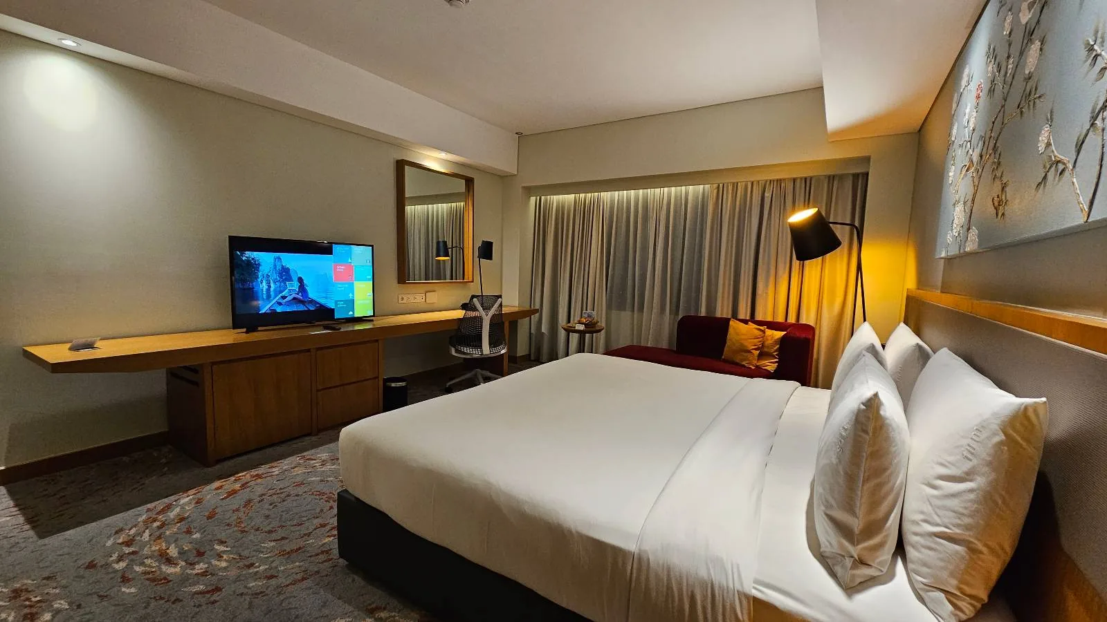 A modern hotel room with a large bed, white linens, a wall-mounted TV, a long desk, a mirror, a sofa, floor lamp, and curtains covering a large window. The room is warmly lit and neatly arranged.