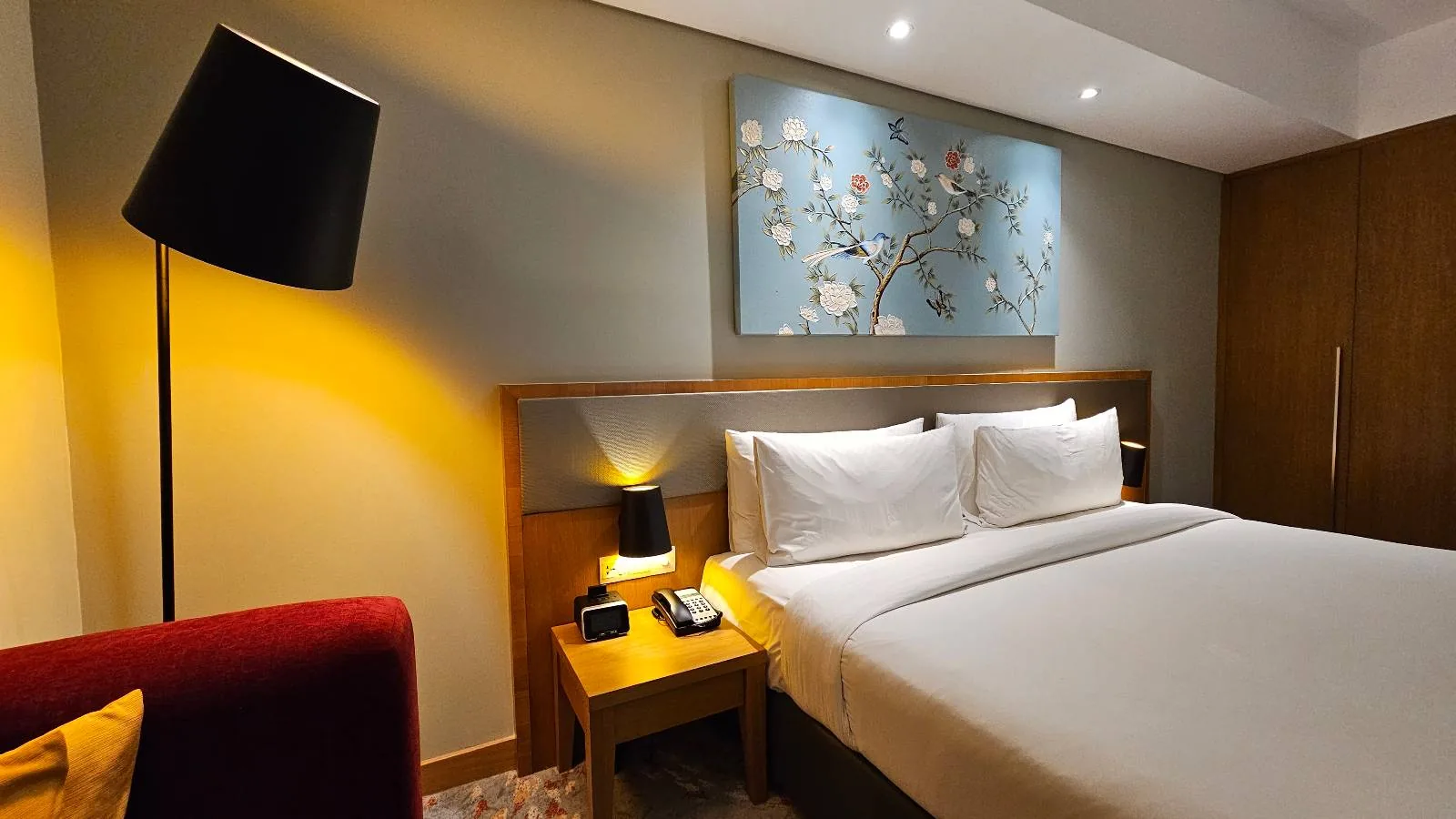 A modern hotel room with a large bed, white linens, two bedside tables with lamps, a wall-mounted floral artwork, a tall floor lamp, and warm lighting.