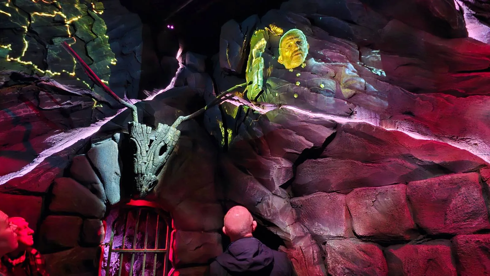 A person stands in front of a rocky wall with a glowing animal skull and bright, colorful images projected onto the surface, creating a mystical and immersive atmosphere.