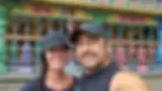 A smiling couple in black caps takes a selfie in front of a colorful, ornate temple with detailed pillars and vibrant patterns.