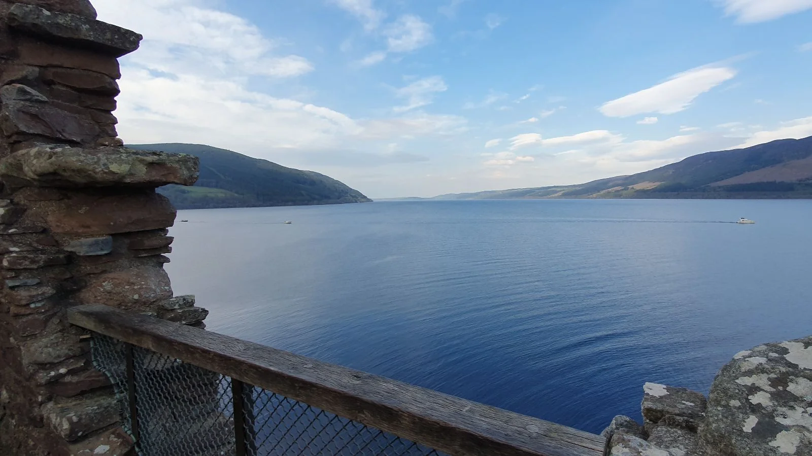 View of Loch Ness from Urquhart Castle, with calm water stretching between rolling hills under a blue sky.