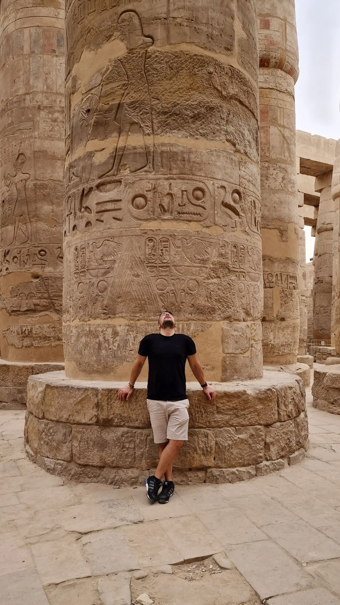 A person in a black shirt and light shorts leans against a massive, ancient stone pillar with intricate carvings. The surroundings include other large columns, suggesting a historical or archaeological site.