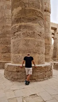 At Karnak Temple, a person leans against one of the mega columns adorned with intricate hieroglyphics and carvings. Clad in a black shirt and beige shorts, they gaze up at the ancient designs, surrounded by other similar mega columns.