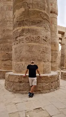 A man in a black shirt and shorts stands looking up at massive ancient stone columns covered in hieroglyphics and carvings during a Luxor day trip to Karnak Temple.