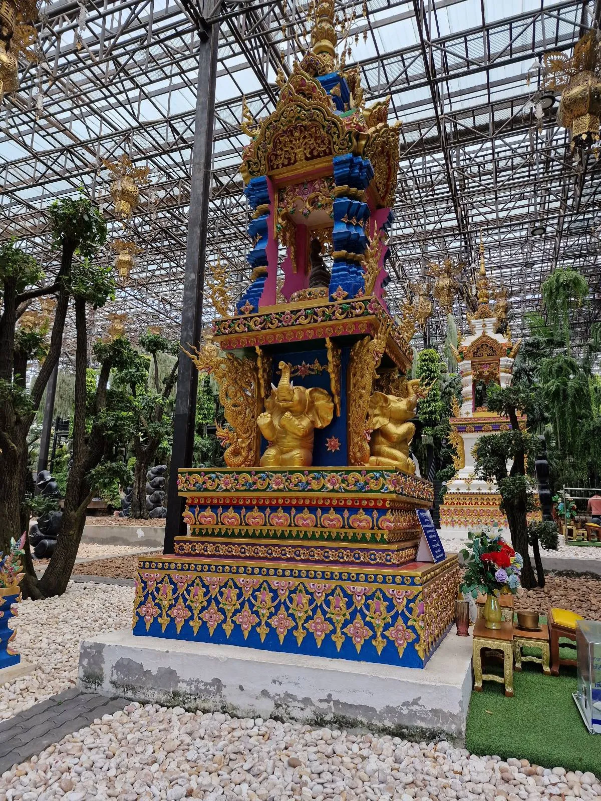 A vibrant, ornate structure sits in a garden, featuring intricate gold and blue decorative patterns. Surrounded by lush greenery and pebbled ground, the structure shows detailed figures and intricate designs under a glass ceiling.