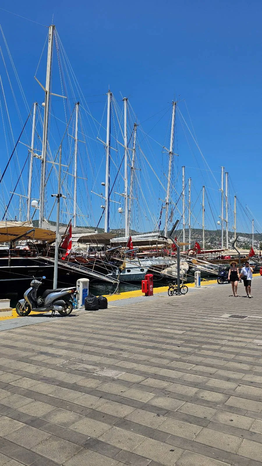Sailboats with tall masts are docked at a marina under a clear blue sky. People walk along the stone-paved waterfront, and several Turkish flags are visible on the boats.
