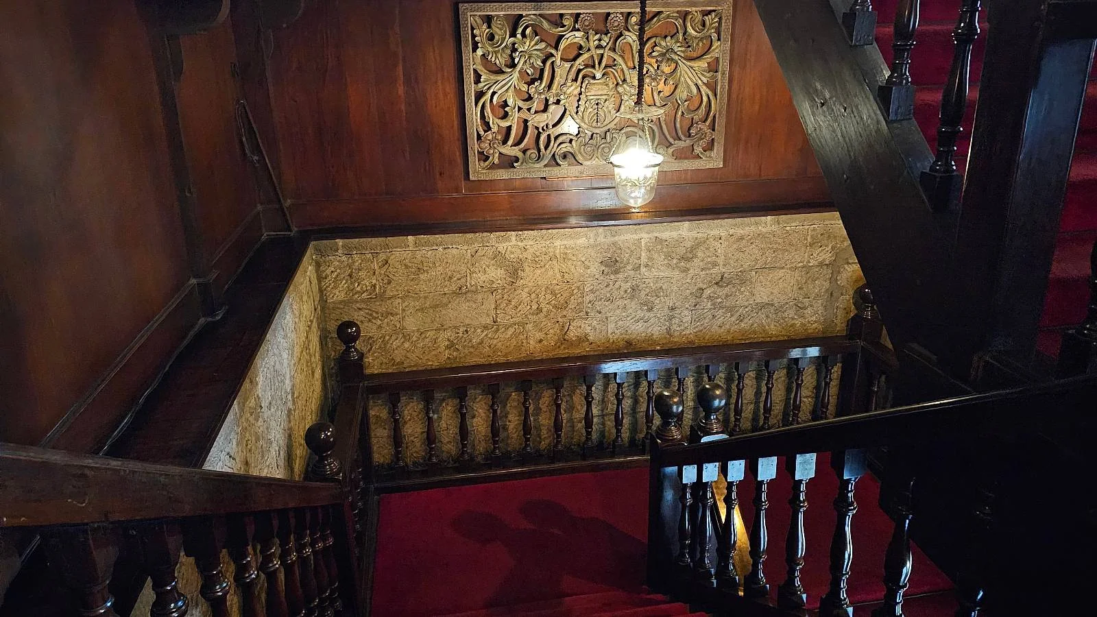 A dimly lit wooden staircase with red carpeting leads down. Ornate carvings decorate the walls, and a hanging light fixture softly illuminates the space. The atmosphere is vintage and elegant.