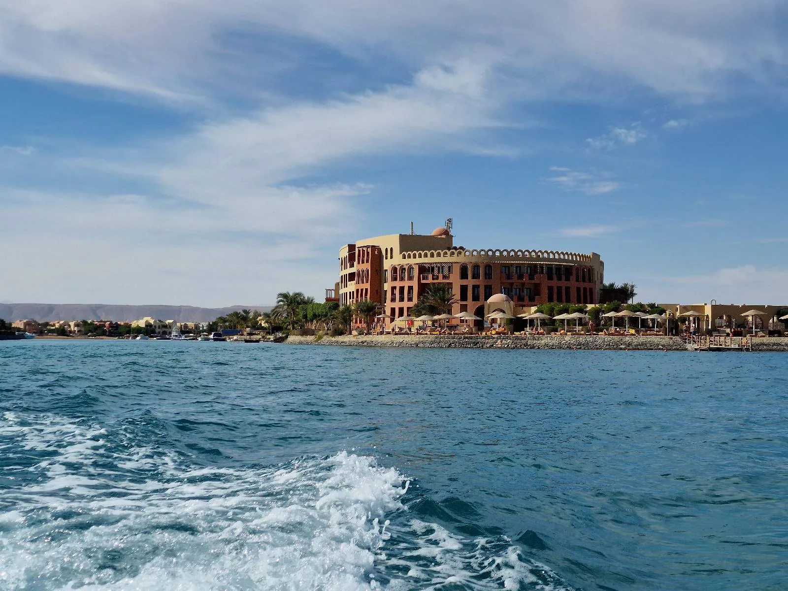 A large, multi-story brick building with arched windows stands on a coastline with clear blue water and a partly cloudy sky in the background. Waves from a moving boat are visible in the foreground.