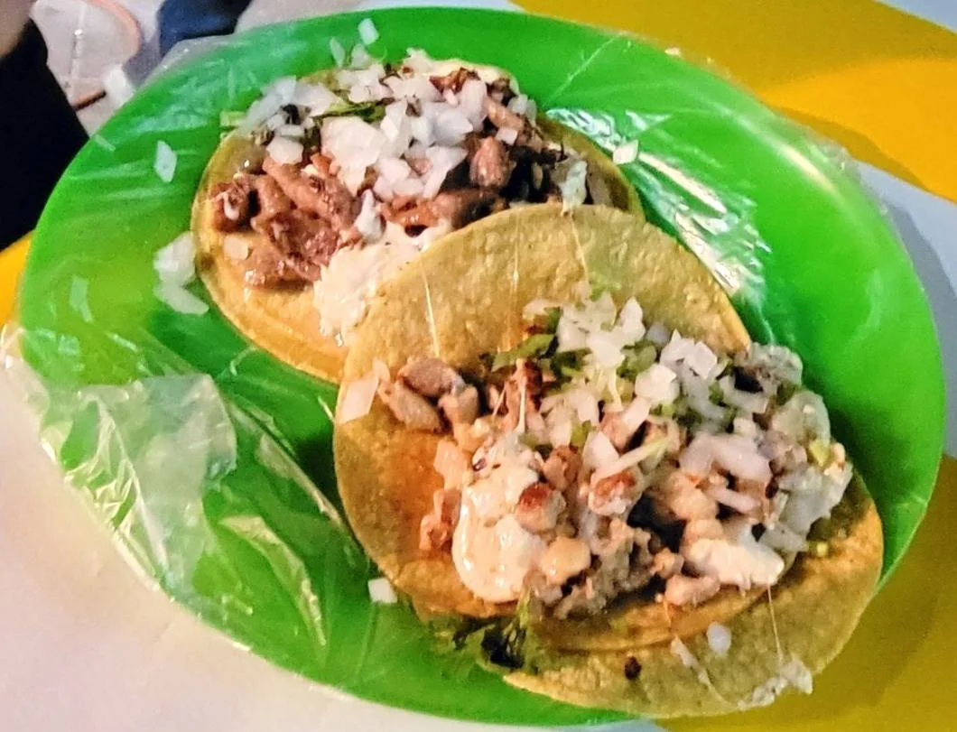 Two tacos on a bright green plate, filled with chopped grilled meat, diced onions, and cilantro. The plate is set on a yellow and white table.