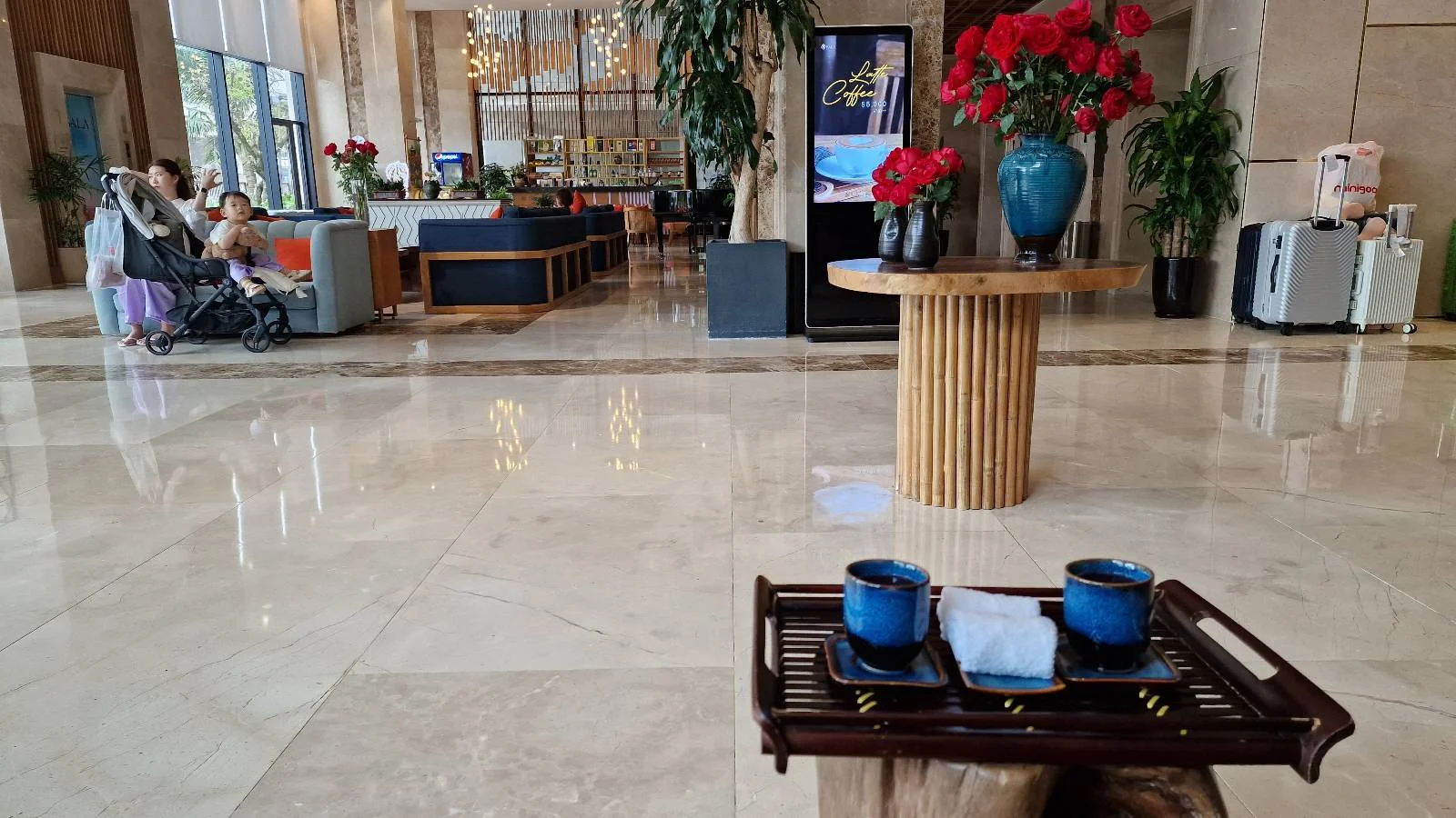 A spacious hotel lobby with a shiny floor, featuring a table with blue cups in the foreground. Red flowers in a blue vase are on a round table. People sit by a window, and a reception desk is visible in the background.