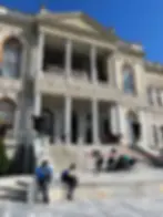 Historic building with ornate facade and columns. People sitting and walking on steps in front. Clear blue sky above.