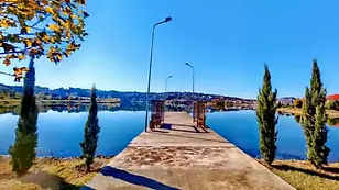 Peaceful lakeside jetty with still blue water reflecting autumn trees and a small town in the distance.