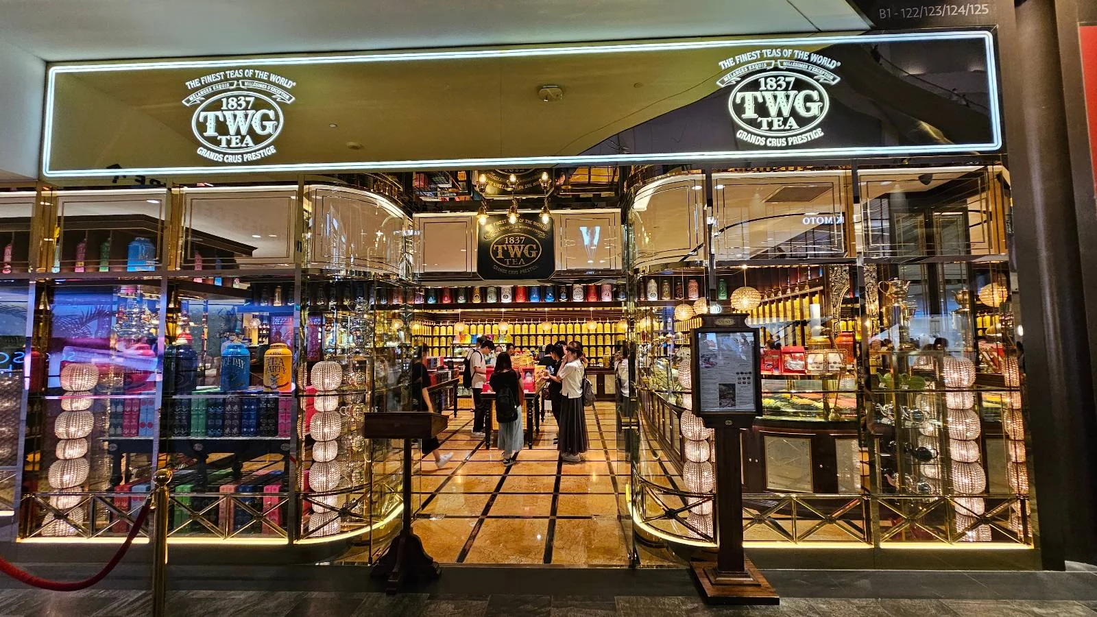 A brightly lit tea store entrance with glass doors. The store's logo is visible above the entrance. Inside, shelves display colorful tea containers. Several people are browsing around. A small menu stand is positioned outside.