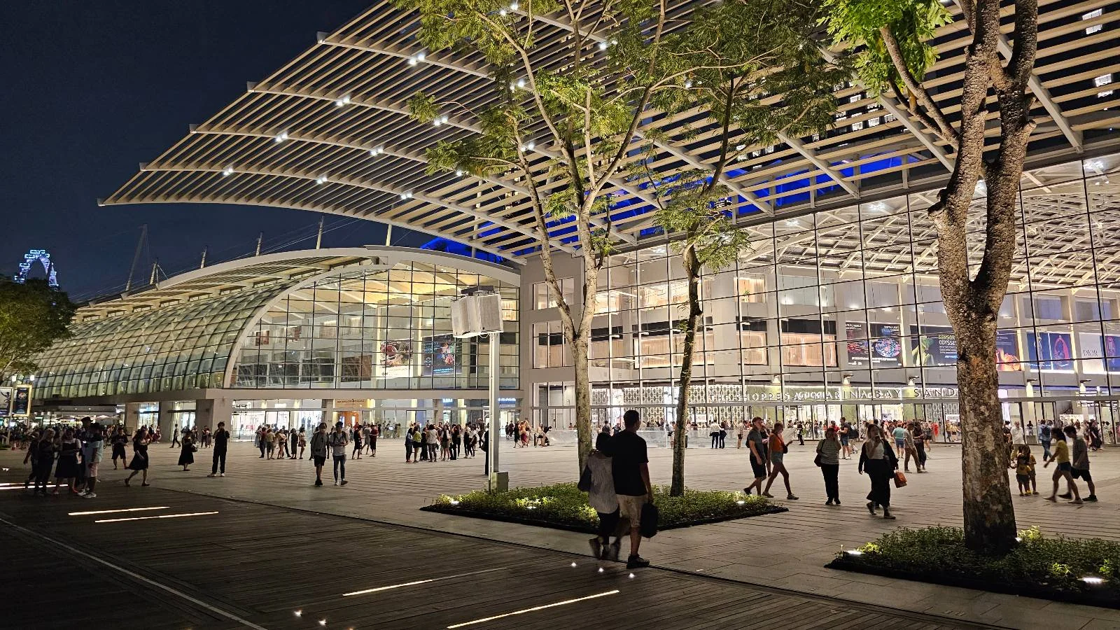 A modern, glass-covered building illuminated at night with people walking nearby. The structure features a unique grid-like roof design and surrounding trees. The night sky enhances the lively and spacious atmosphere.