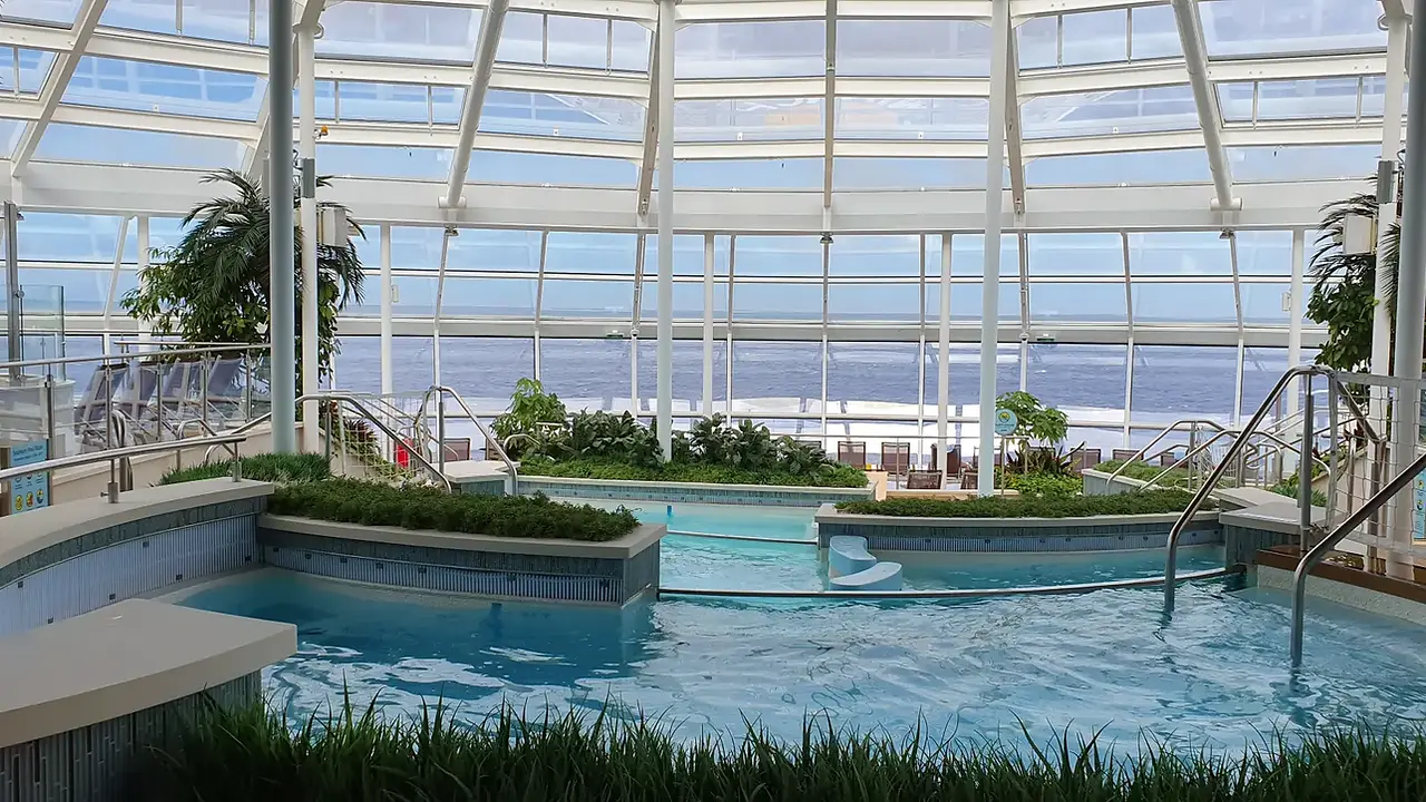 The glass-roofed indoor pool area aboard Ovation of the Seas with greenery and ocean views.