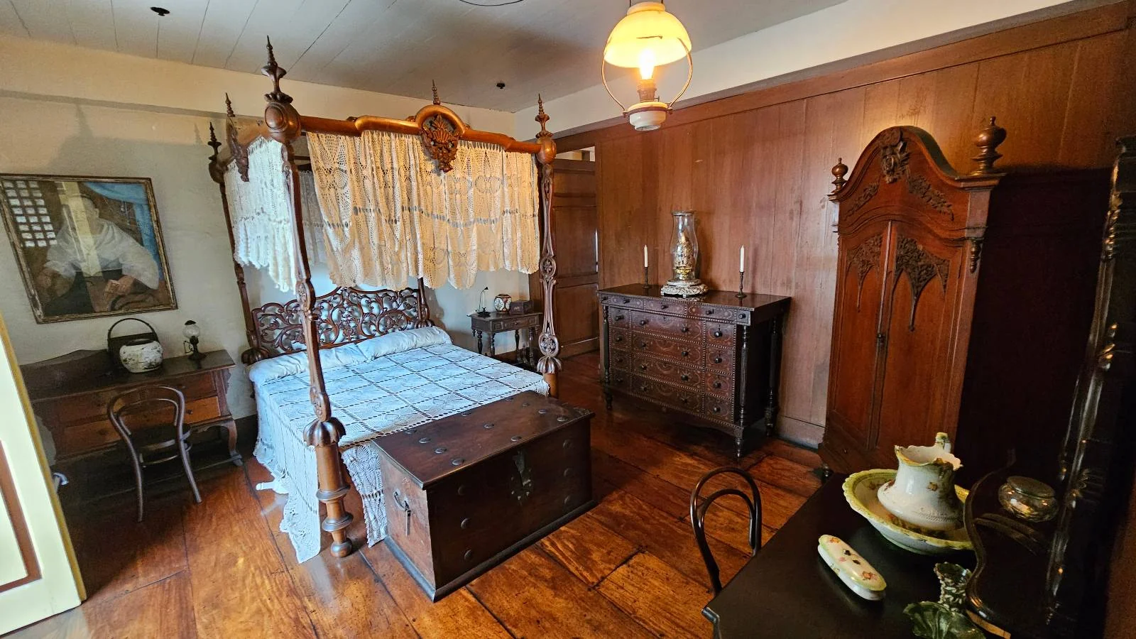 A vintage bedroom with a wooden four-poster canopy bed, ornate dresser, wardrobe, and bedside tables. The room features wooden paneling, soft lighting, and classic decor, including a decorative jug and basin on a table in the foreground.