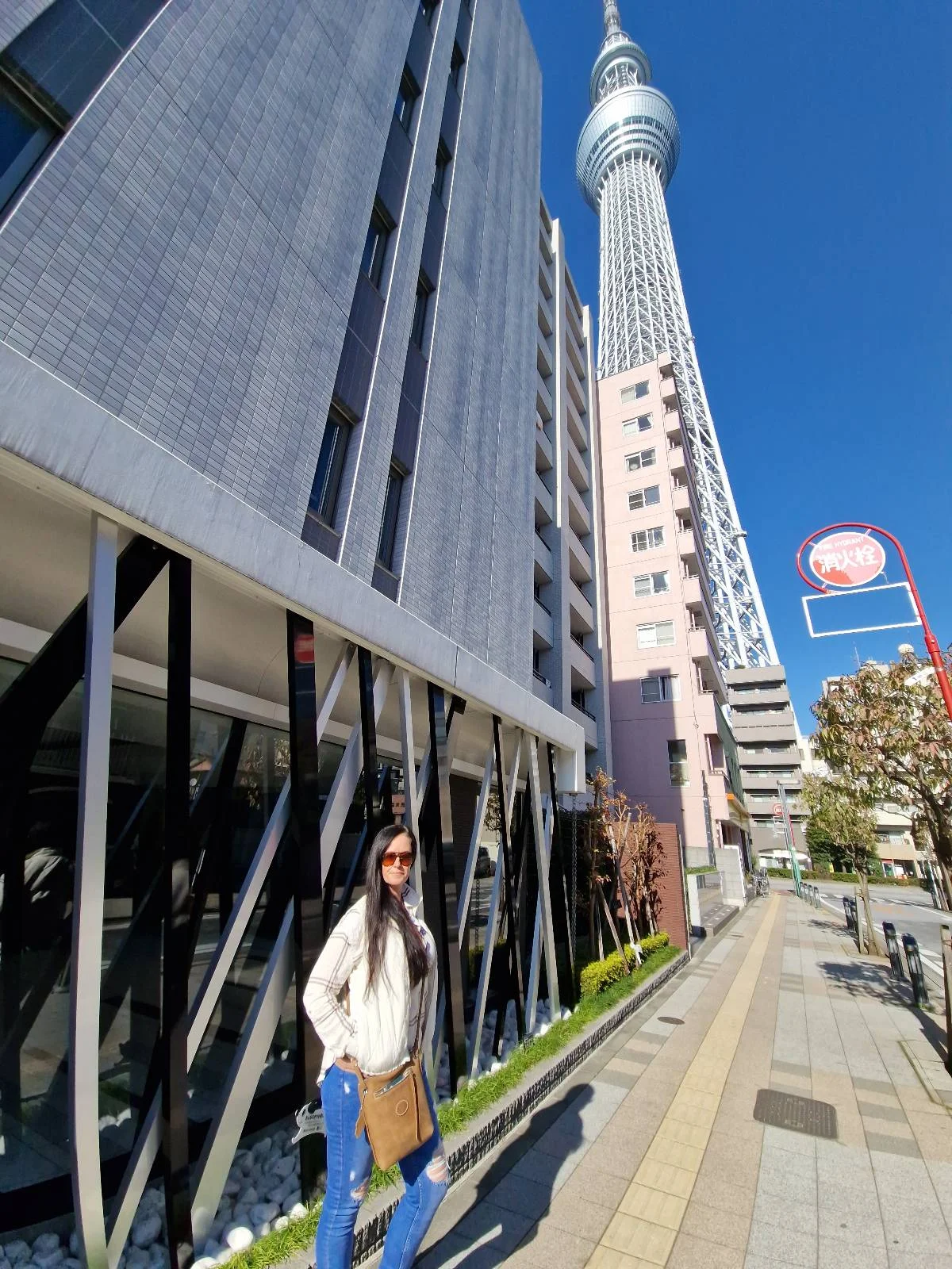 A person stands on a sunny sidewalk wearing sunglasses, a white jacket, and blue pants. Behind them is a tall, modern building with the Tokyo Skytree visible in the background under a clear blue sky.