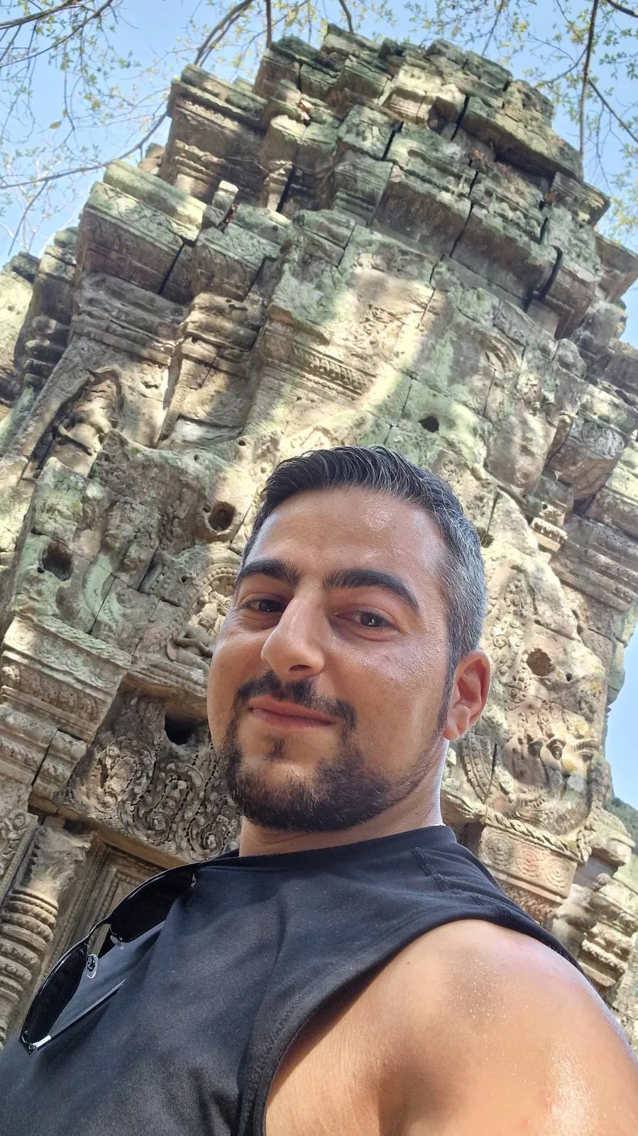 A man with short dark hair and a beard takes a selfie in front of an ancient stone temple with intricate carvings, under a clear blue sky.