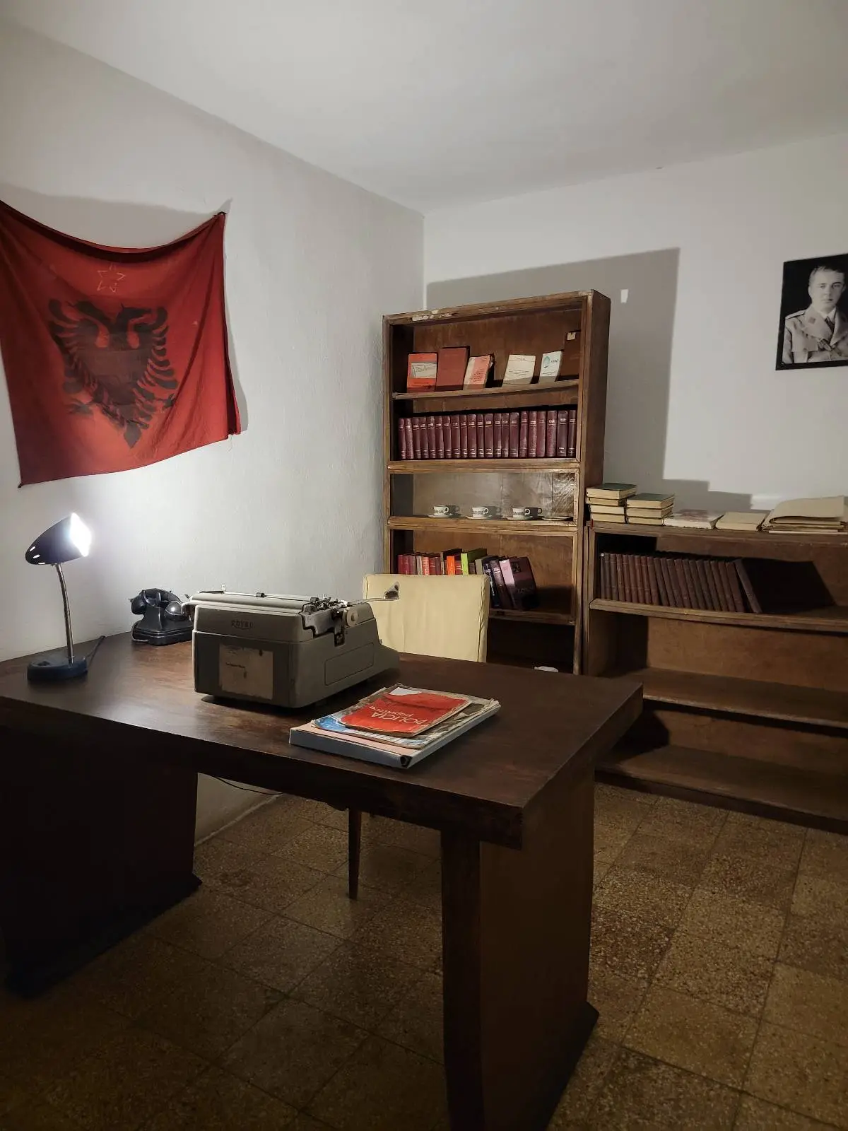 A small office with a desk, lamp, books, and files. An Albanian flag hangs on the wall, and bookshelves hold documents. A black-and-white portrait is displayed above a cabinet.