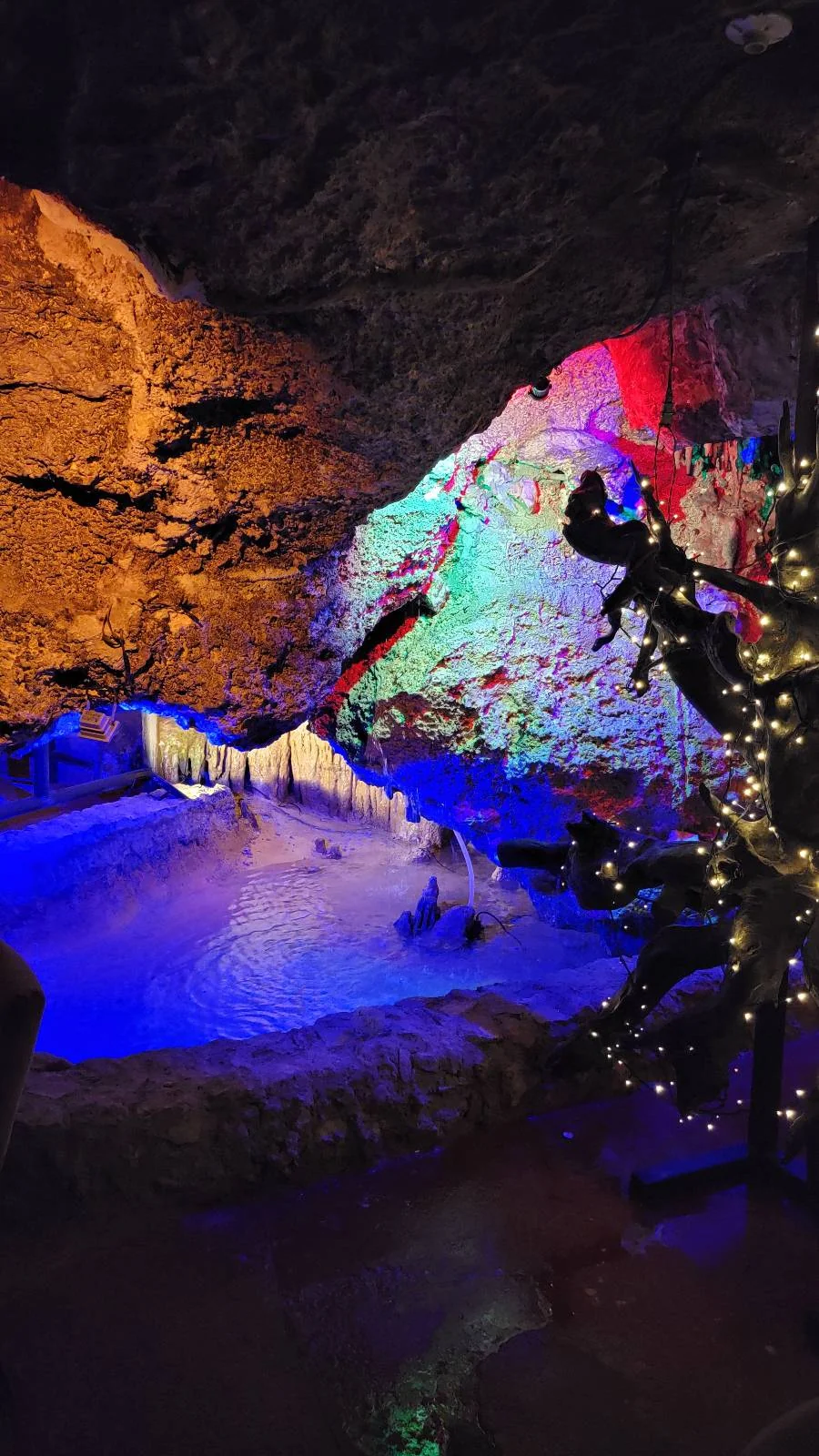 Cave interior with illuminated rock formations in blue and red light. A small pool reflects the light. A decorated tree with string lights is on the right, adding a festive touch to the scene.