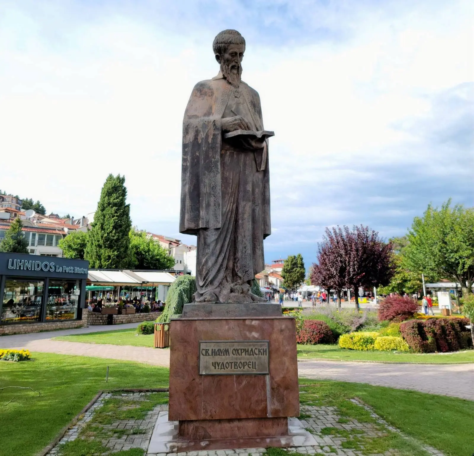 A tall statue of a bearded figure holding a book, standing on a pedestal in a park setting. The pedestal has an inscription. Surrounding the statue are green lawns, a paved path, and trees. A market stall is visible in the background.