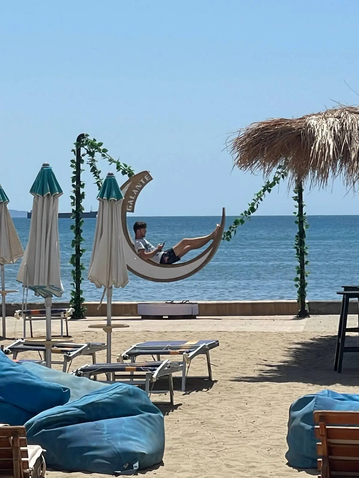 Person relaxing in a crescent moon-shaped swing by the beach, with umbrellas, lounge chairs, and the sea visible in the background under a clear blue sky.