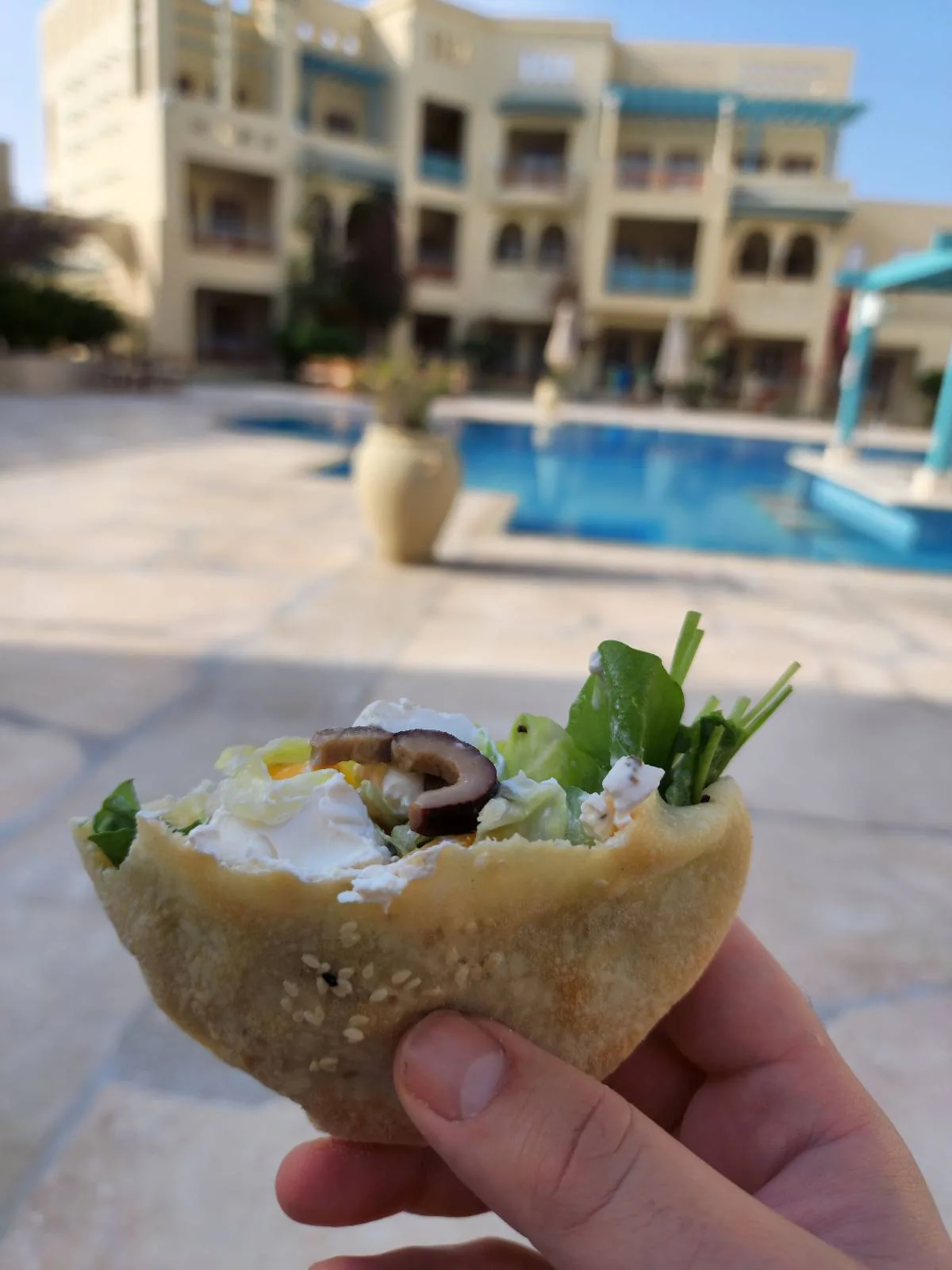 A hand holds a pita bread sandwich filled with vegetables and olives, with a large swimming pool and residential buildings in the background.