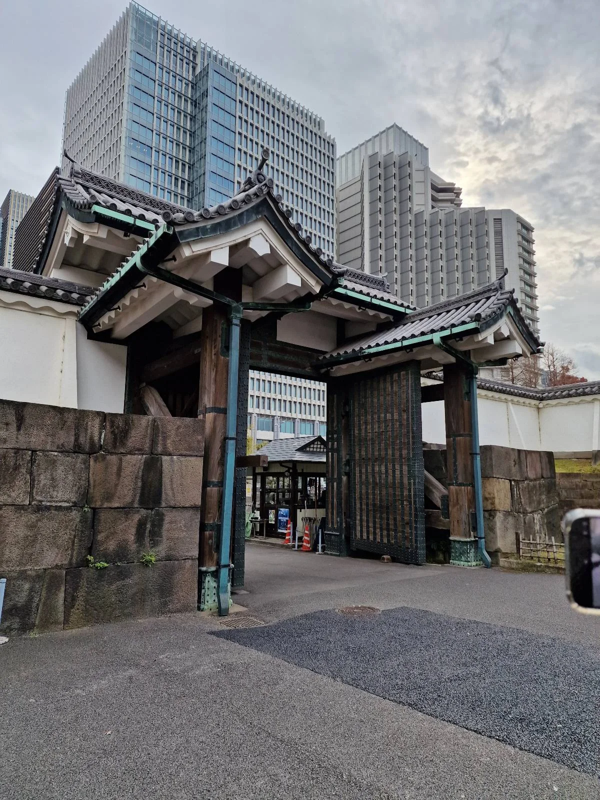 A traditional Japanese gate, characterized by its wooden structure and tiled roof, stands in contrast to modern skyscrapers in the background. The sky is overcast, adding a muted tone to the scene.