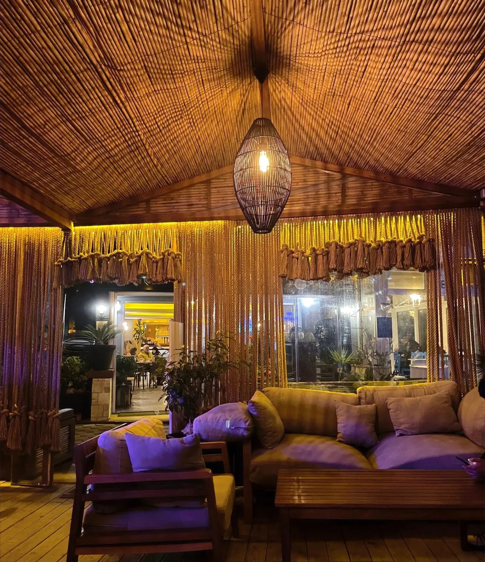 A cozy indoor lounge with wooden furniture, soft cushions, warm lighting, and a woven ceiling. A pendant lamp hangs above a sofa set, creating a relaxed and inviting atmosphere.