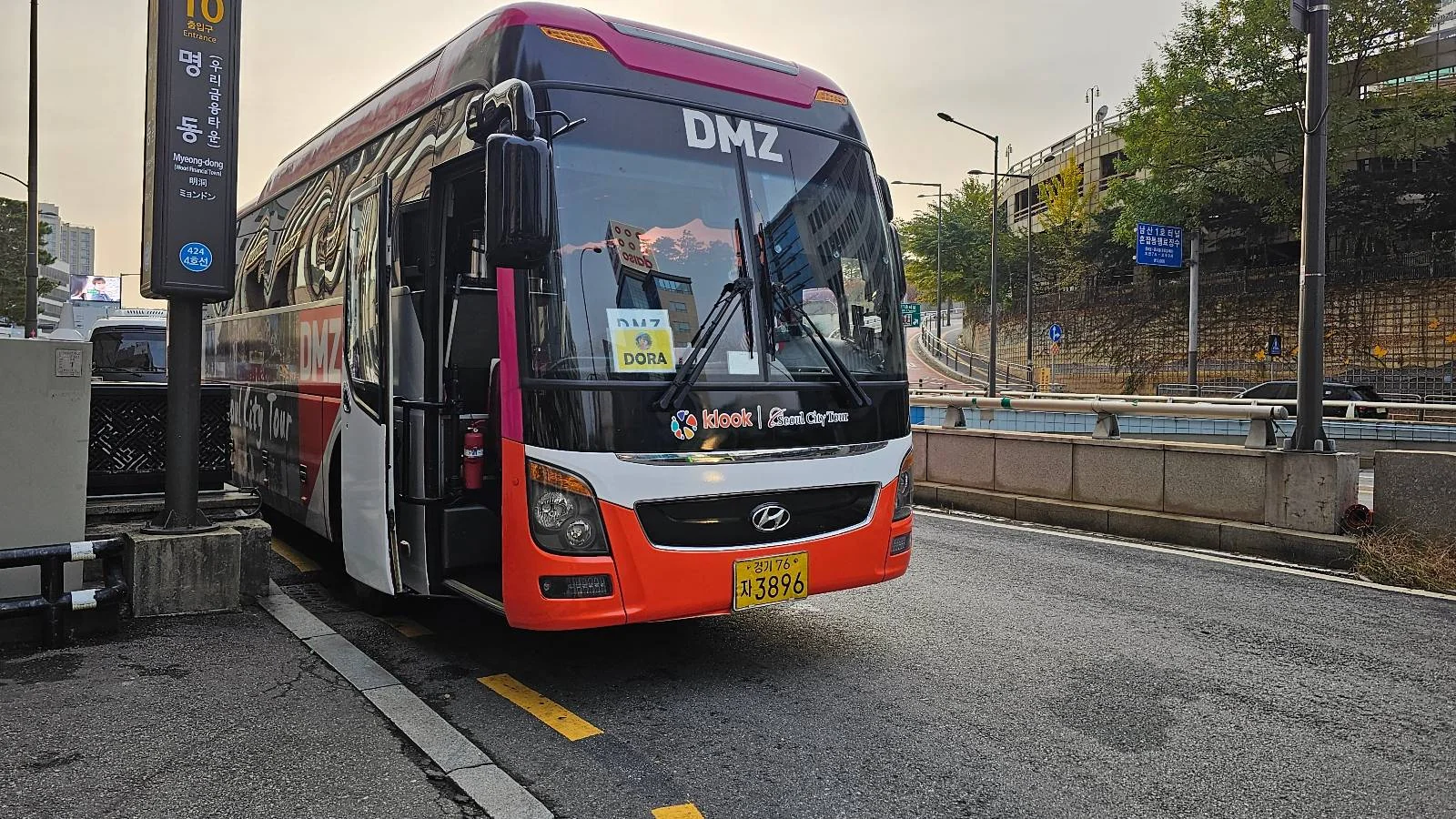 A red and white bus labeled "DMZ" is parked at a bus stop on a city street. The front displays a sign with Korean writing. There are trees and road signs in the background under a cloudy sky.