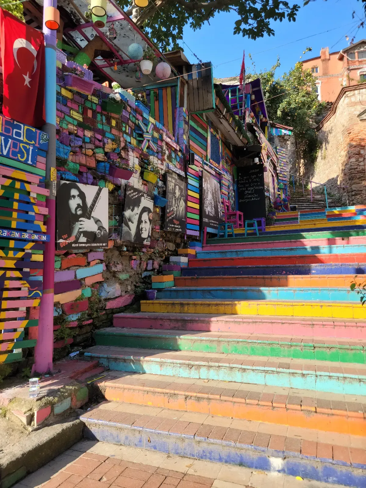 Colorful outdoor stairs decorated with bright, painted patterns and stripes, alongside a vibrant wall covered in mosaics, graffiti, flags, and artistic portraits, under a clear blue sky.