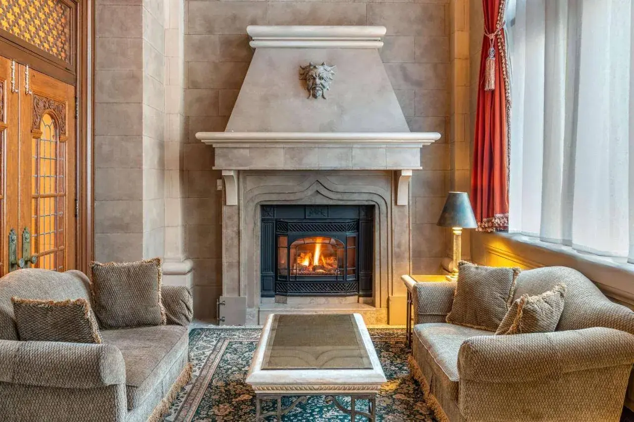 A cozy living room features a stone fireplace with a lit fire, flanked by two beige sofas with cushions. A coffee table is in the center on a patterned rug. Large windows with red curtains let in natural light, and a lamp is beside one sofa.