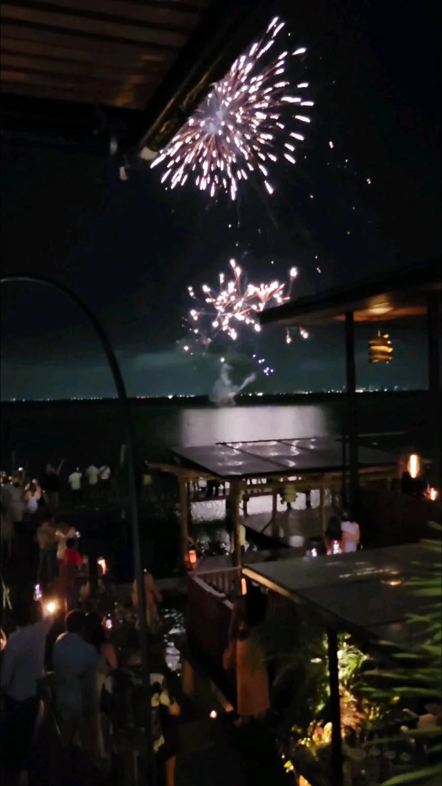Fireworks light up the night sky over a waterfront. People gather on an outdoor terrace, illuminated by warm lights, watching the display. A body of water is visible in the background, reflecting the fireworks.