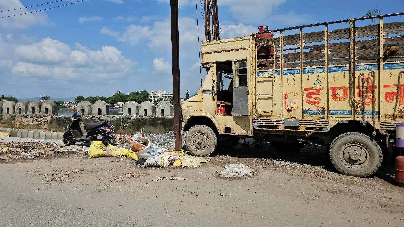 A yellow truck is parked beside a road with construction debris and garbage bags scattered nearby; a motorcycle is parked next to the debris and several concrete structures are visible in the background under a blue sky.