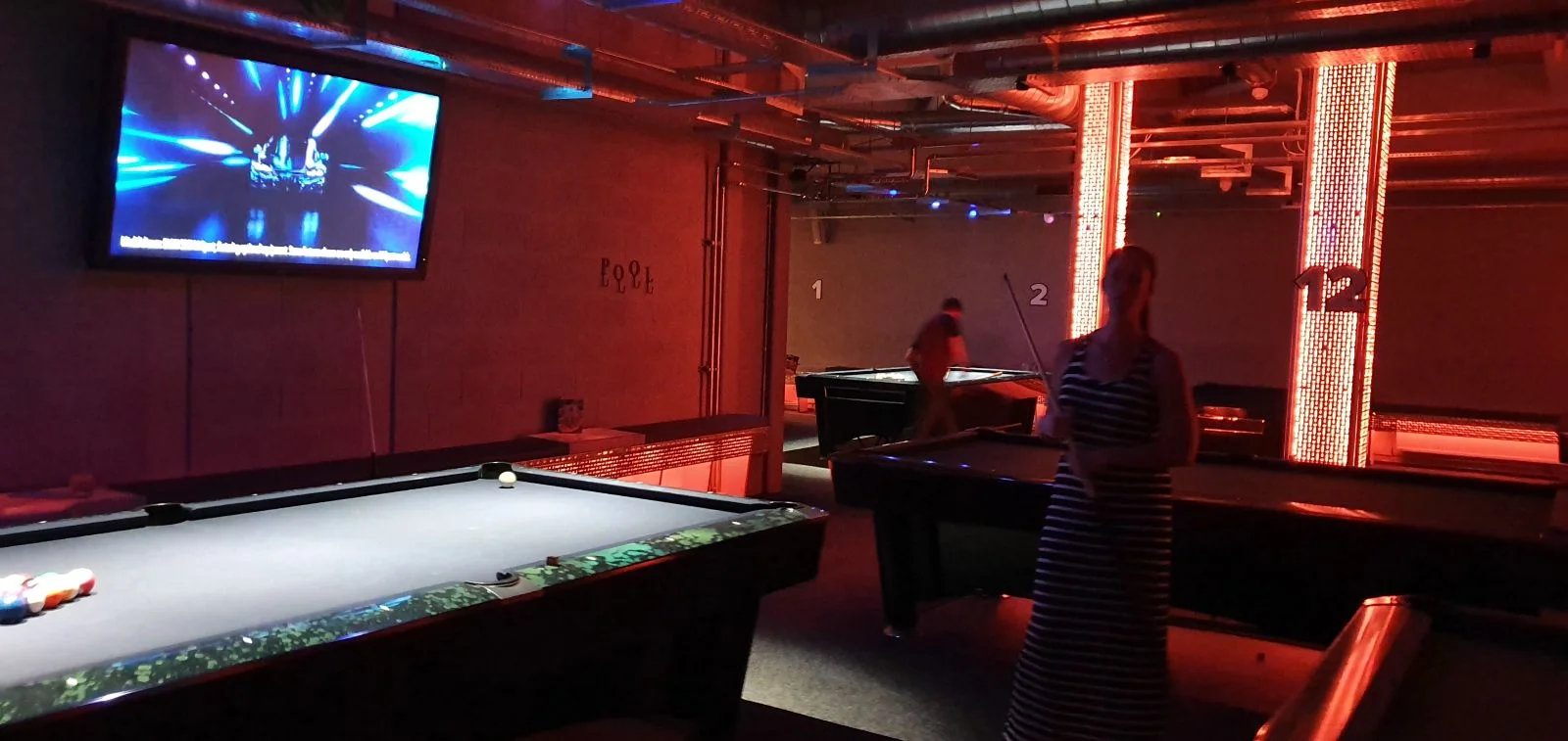 Dimly lit billiard room with pool tables. A person plays near a neon-lit pillar labeled "12." A screen displays a blue-lit stage.
