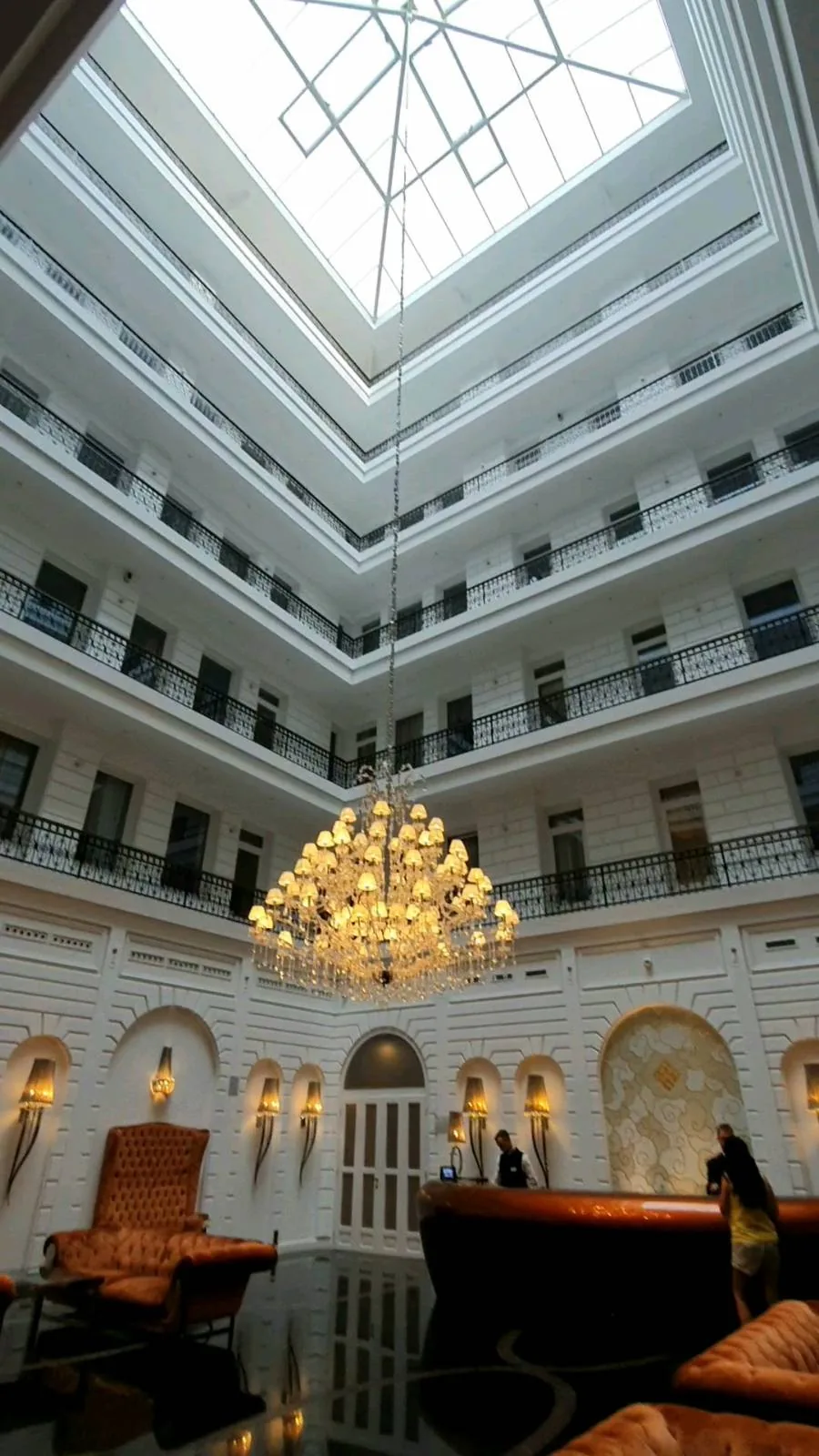 A grand hotel lobby with a large, ornate chandelier hanging from a high, glass-paneled ceiling. The space is surrounded by multiple stories of balconies, and the floor reflects the light. Two people stand at a dark reception desk.