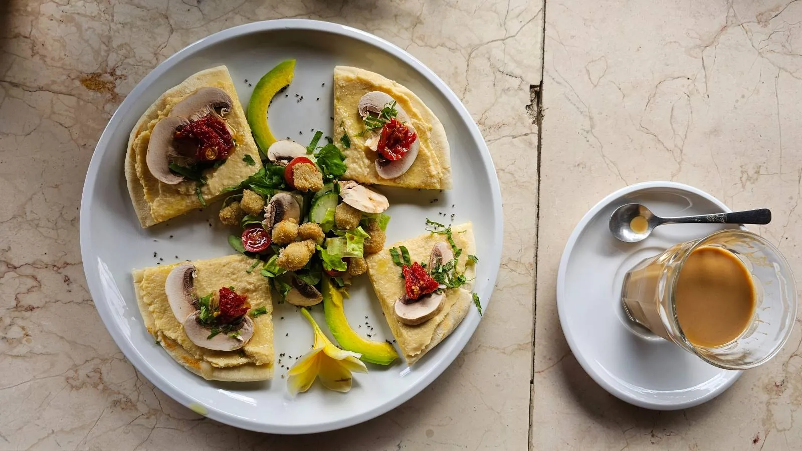 A plate with four triangle-shaped pieces of food topped with sliced mushrooms and red garnish, alongside a serving of mixed vegetables and herbs. A small cup of espresso with a spoon rests on a saucer nearby.