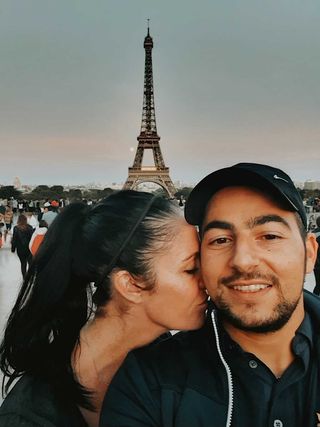 A couple taking a selfie with the Eiffel Tower in the background. The woman is kissing the man's cheek.